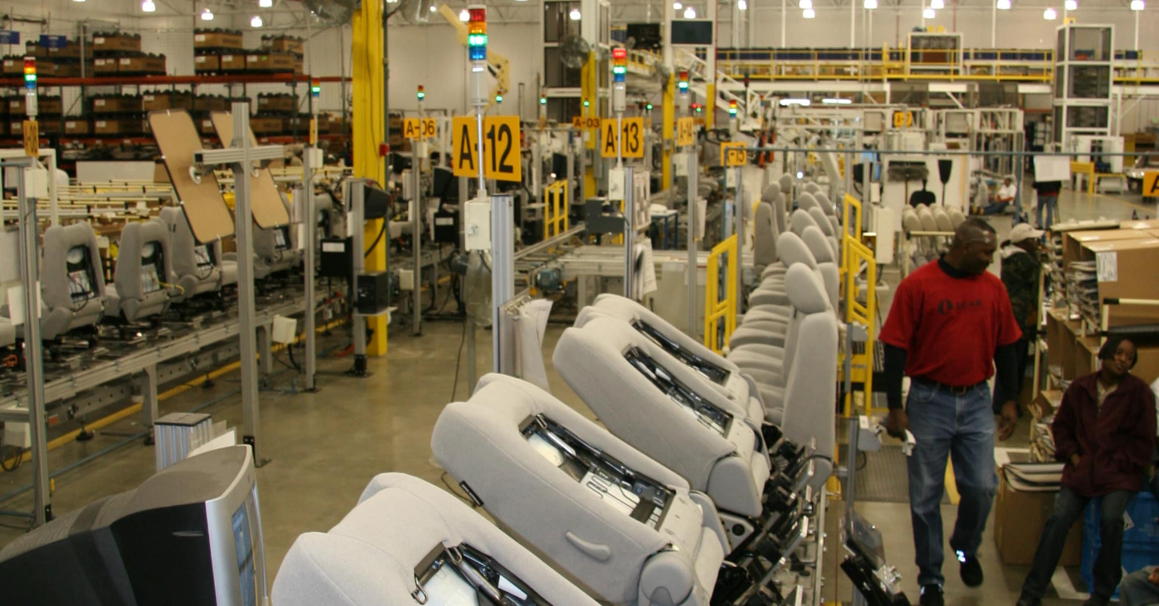 Factory assembly line with rows of gray reclining chairs and workers moving between stations.